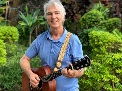 A smiling man in a blue shirt is standing outside and holding a guitar.