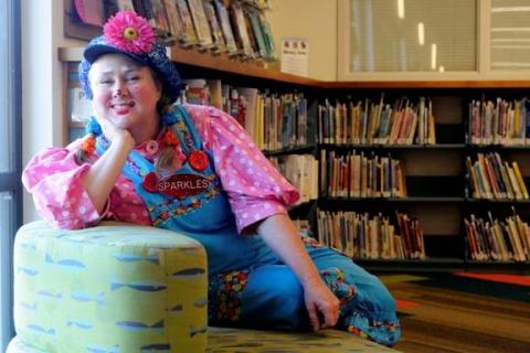 A woman with a friendly smile who is dressed as a clown is sitting in front of library book shelves.