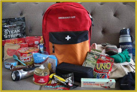 A red emergency backpack with its supplies displayed around it including dried food, water, flashlight, and a card game.