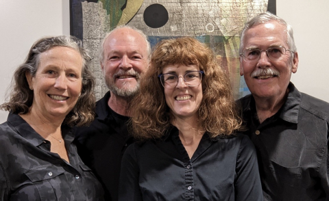 Four smiling adults posing together indoors against an abstract art background.