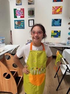 Young girl wearing a bright yellow apron is standing in an art studio, smiling and ready to create art!