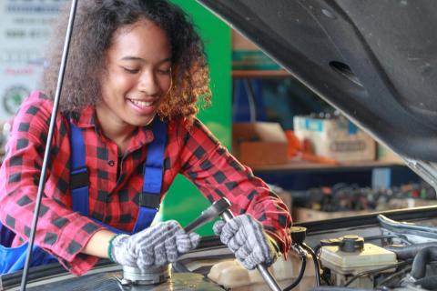 Female presenting individual wearing gloves is looking at and working on a car engine using tools.