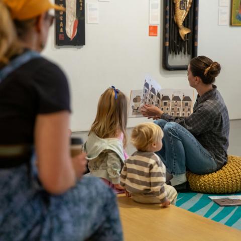 A family is watching a storytime on a bright colored rug in the Mingei International Museum.