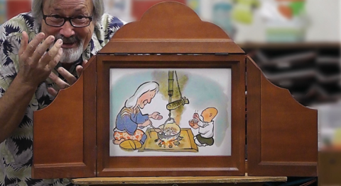 A man with gray hair, a beard, and glasses, stands next to a wooden theater with a cute illustration of a grandmother and grandson eating.