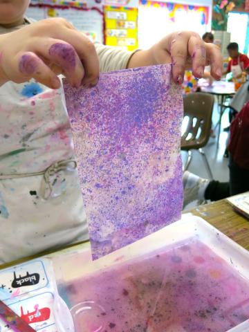 Picture of a child holding up a piece of paper they made a marble pattern on using purple chalk.