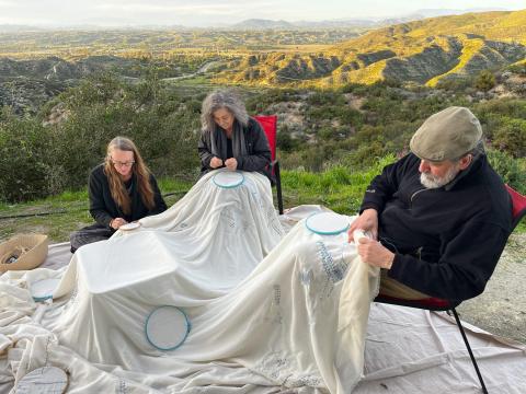  3 people sit outside against a valley landscape embroidering the same piece of cloth with hashmarks that  are significant to them.  