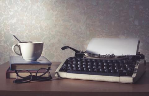 Desk with a typewriter, eyeglasses, and a cup on top of books.