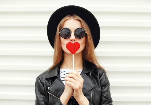 Young person wearing a hat and sunglasses holding a heart-shaped lollipop in front of their mouth