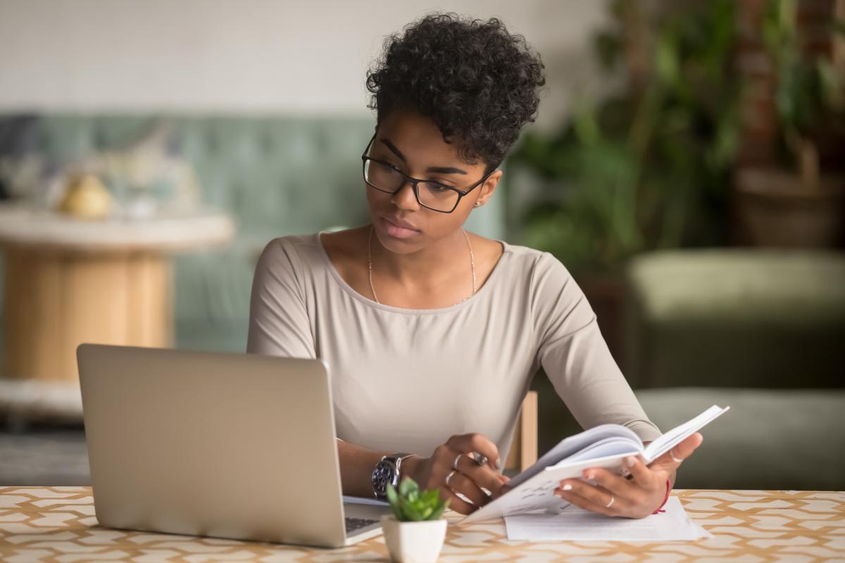 A black woman looks at an open laptop while holding an open book.