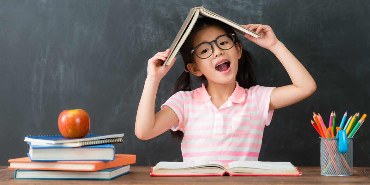 Little girl wearing glasses sitting at a desk holding a book on her head and smiling