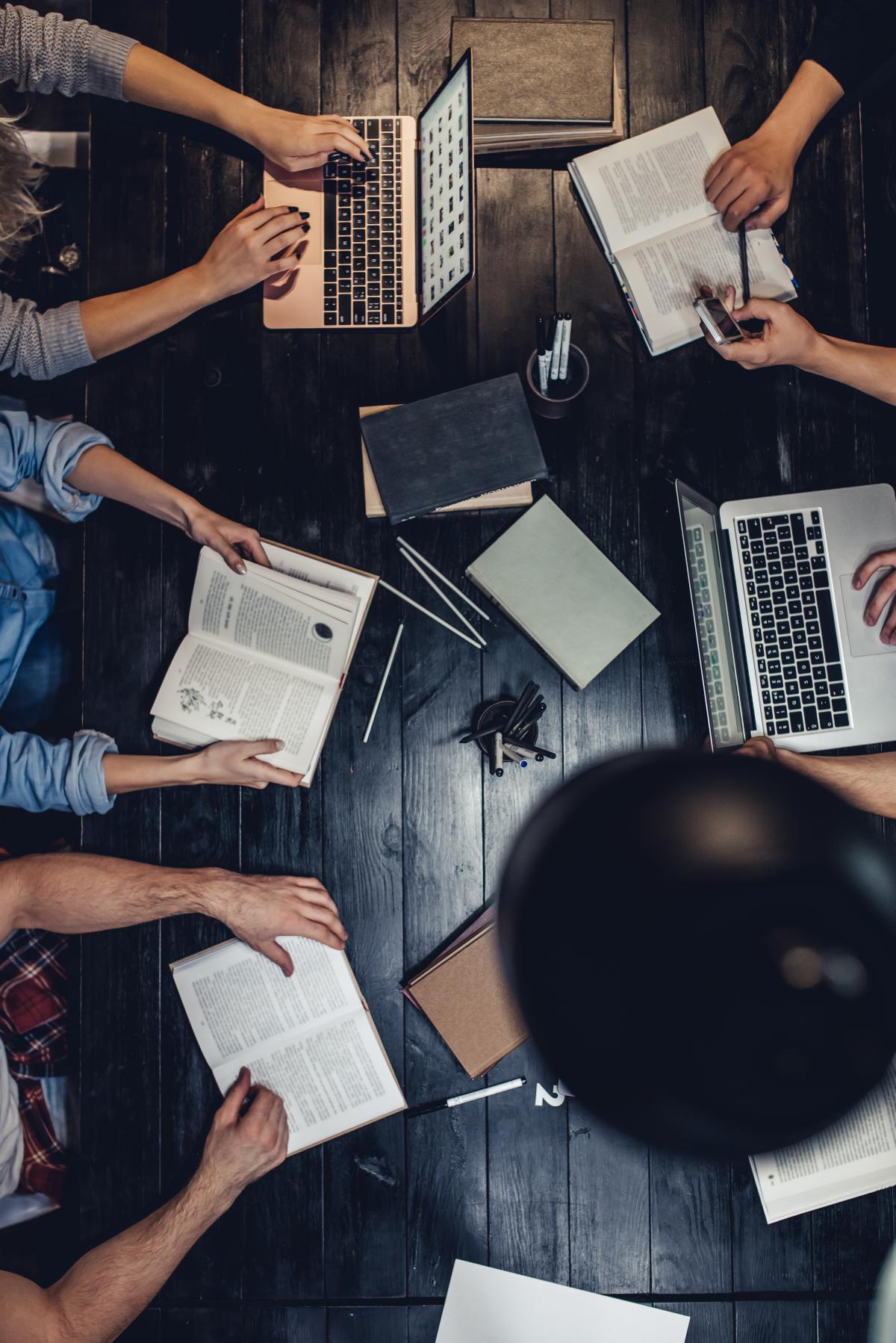 An overhead view of 6 people sitting at a table with laptops and study materials