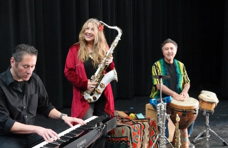 Group photo of a man playing keyboard, a woman with a saxophone, and a man playing drums.  