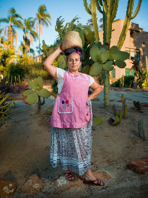 Alt Text: Photographic portrait of a woman holding a basket on her head, standing in front of cactuses by artist John Raymond Mireles.
