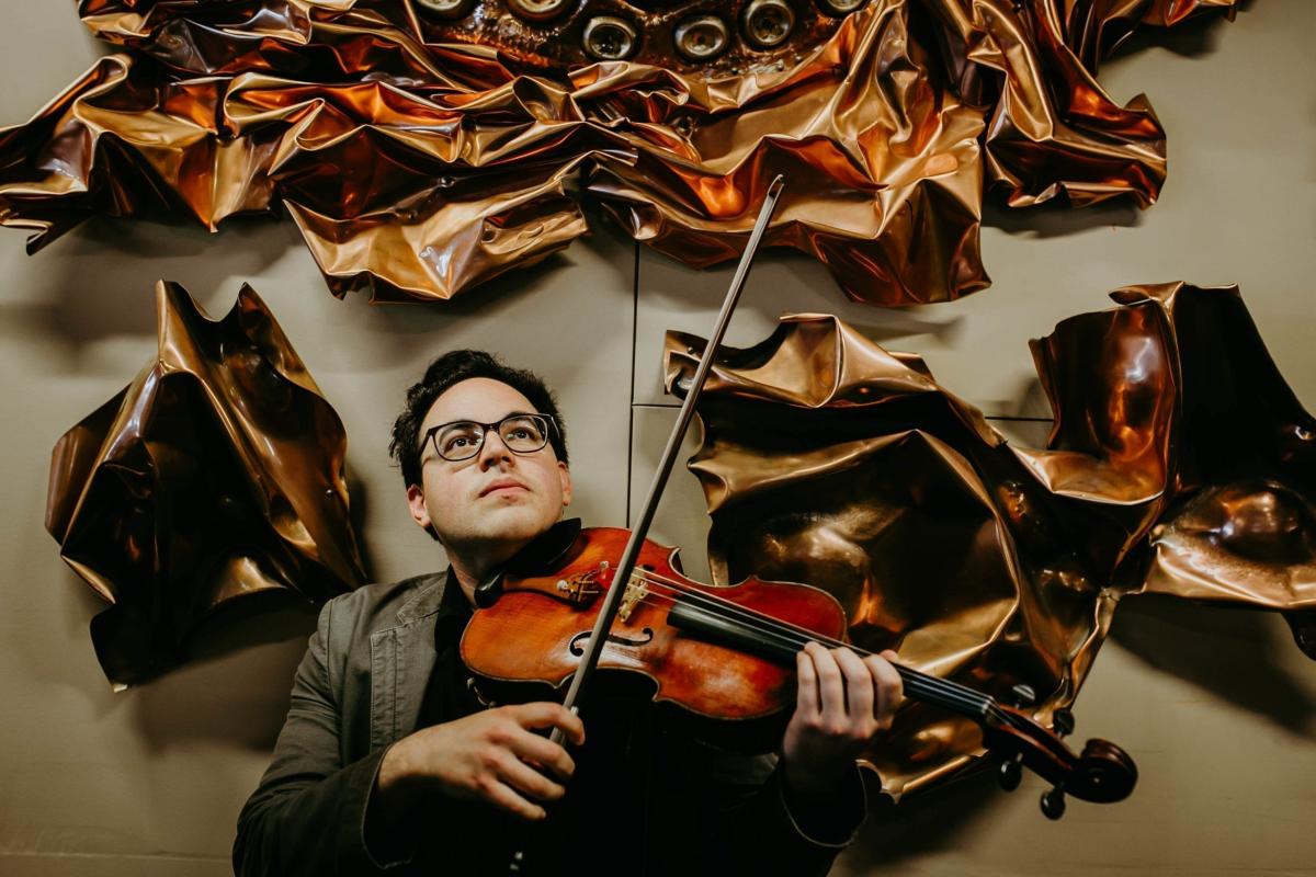 Photo of dark haired man with glasses holding a violin surrounded by wavy metal art