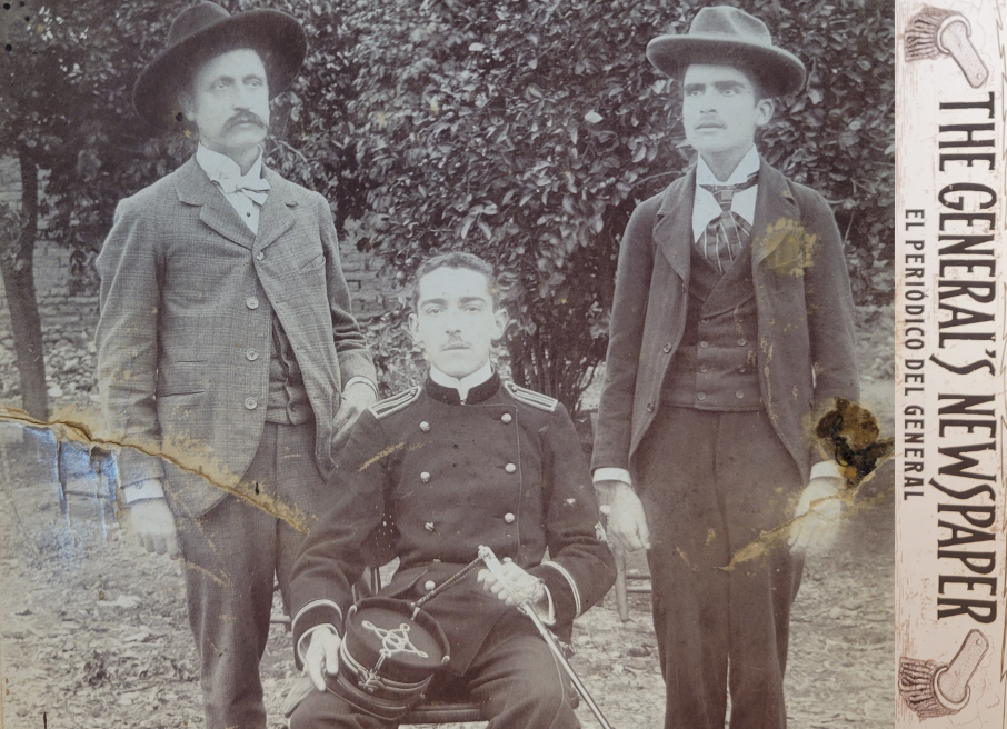 Historical photograph of three men, two standing, one sitting wearing an army uniform with the words “The General’s Newspaper, El periódico del general” along the side.