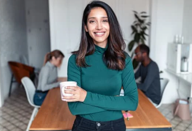 Woman with coffee cup stands in an office with people and a desk behind her. 