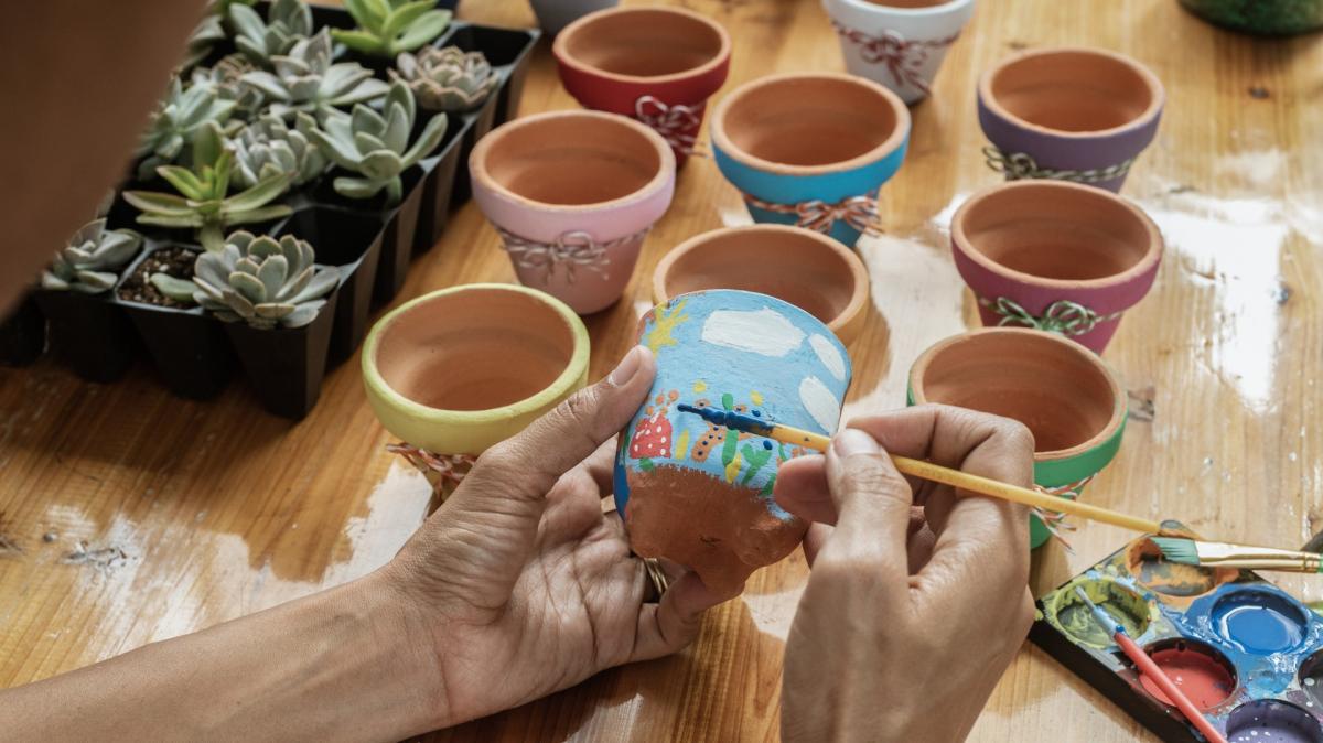 Hands painting a small terra cotta pot with pots and plants on the table in the background