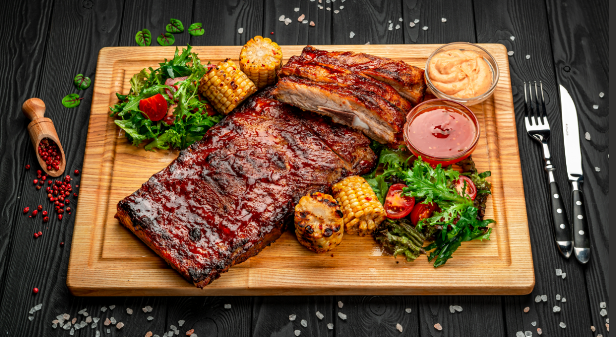Photograph of cooked ribs, corn, salad, and two dipping sauces on a cutting board.