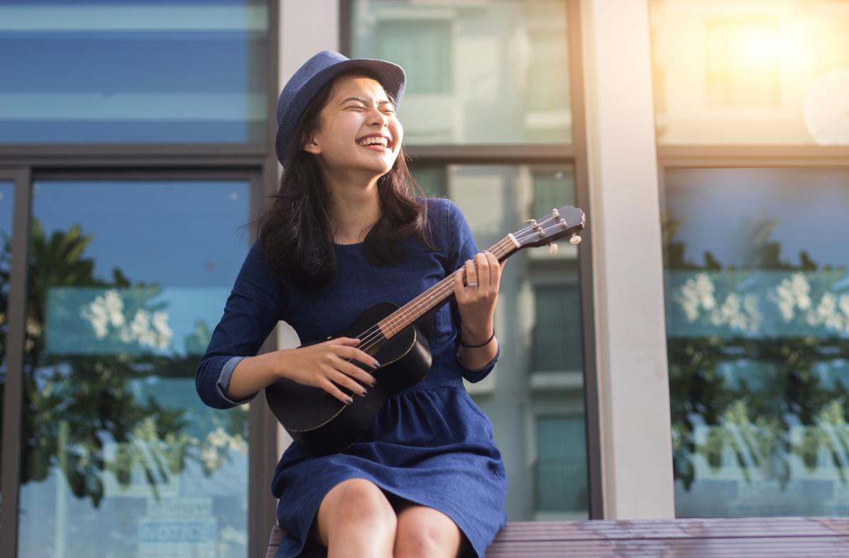 Photo of a female-presenting person wearing a blue dress and hat, holding a ukulele, and smiling