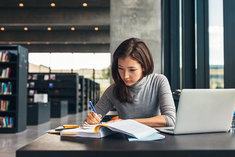 woman studying with book, laptop, notes in library setting